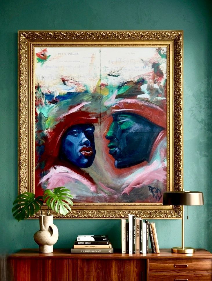 Colorful abstract portrait in a gold frame above a wooden console table with books, lamp, vase, and plant.
