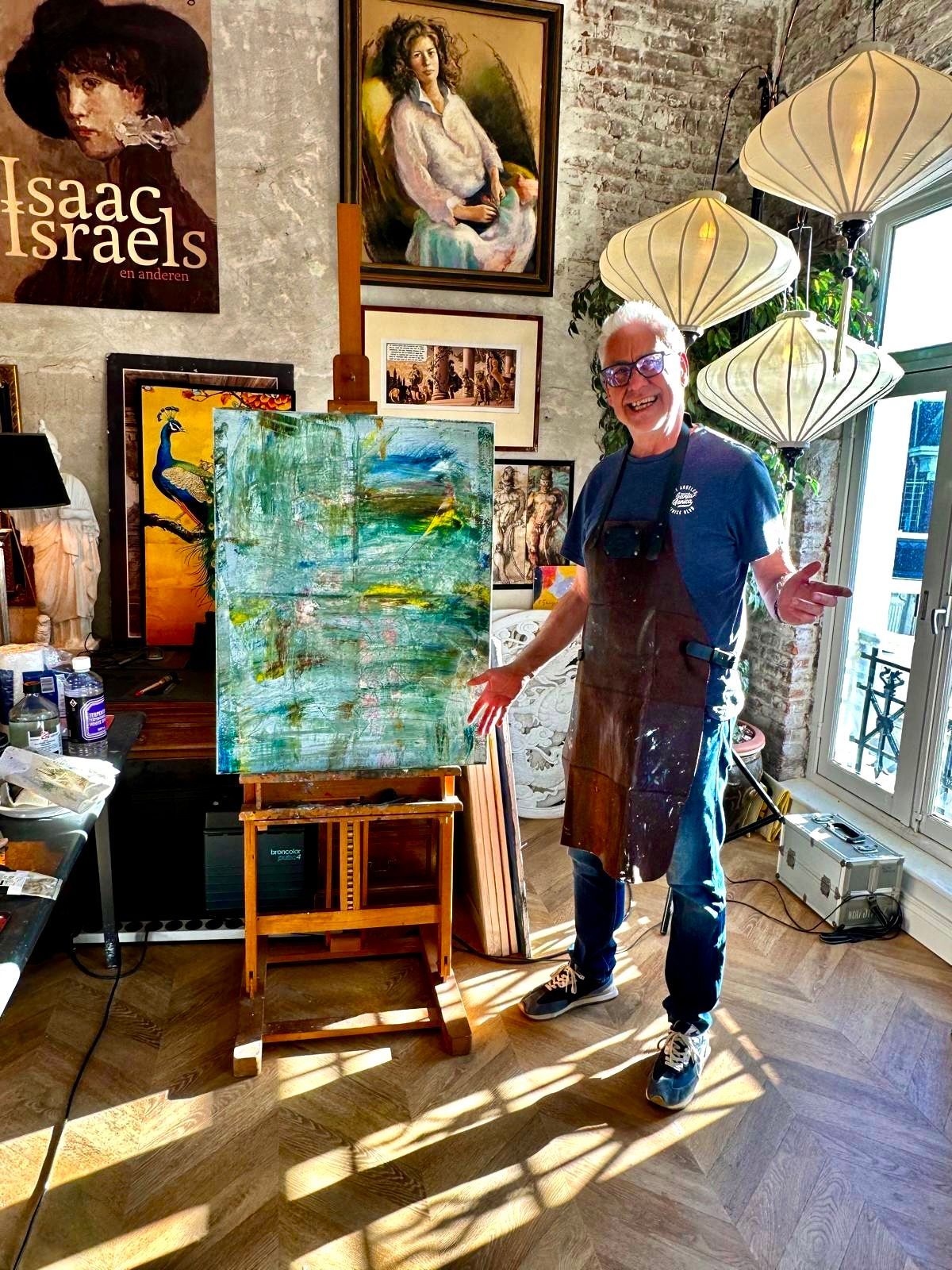 Kunstenaar staat naast een abstract schilderij op een ezel in een zonovergoten atelier met ingelijste kunstwerken aan de muur.