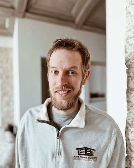 A man with a beard wearing a gray sweatshirt smiles in a bright, indoor setting.