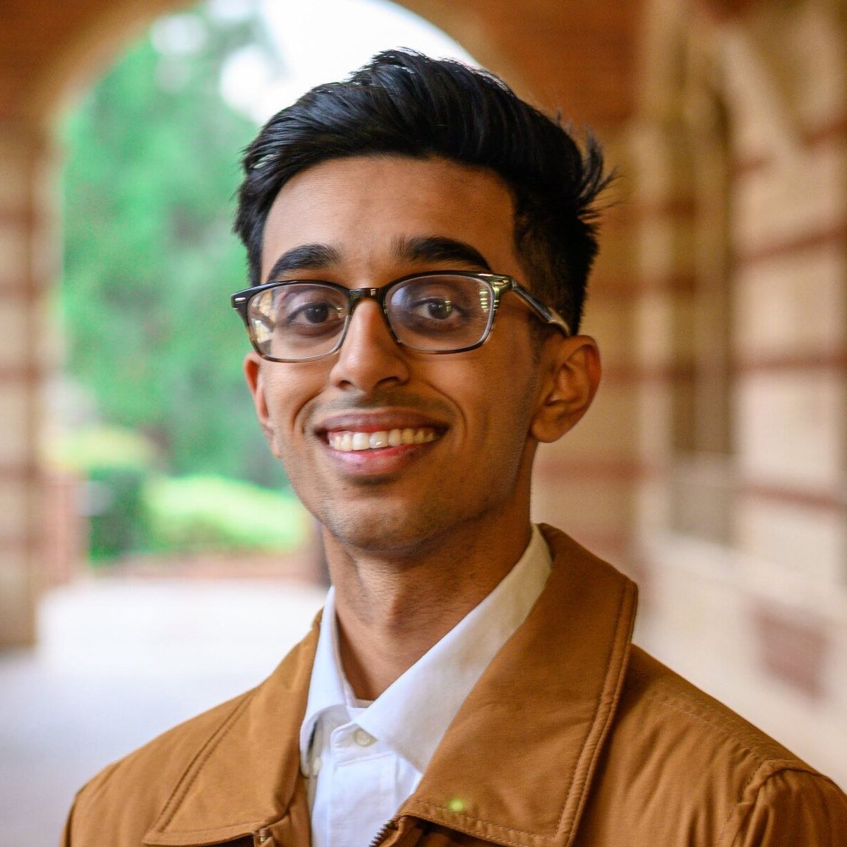 A young man wearing glasses and a brown jacket is smiling for the camera.