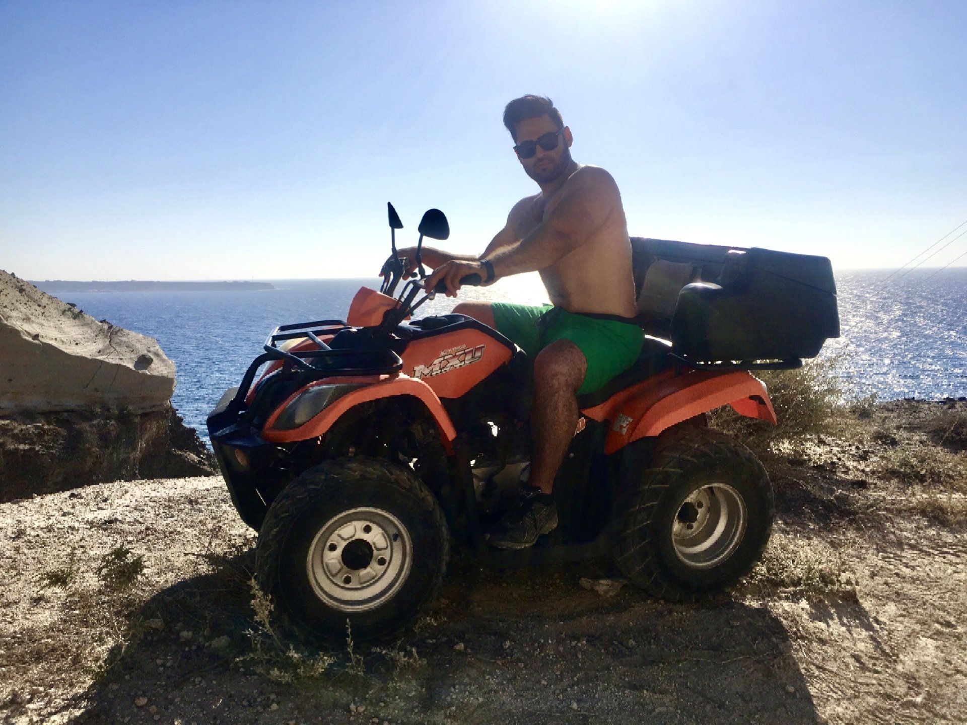 A man without a shirt is sitting on an atv near the ocean