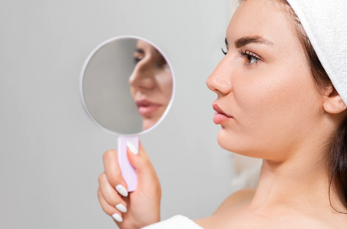 A woman with a towel on her head is looking at her face in a mirror.