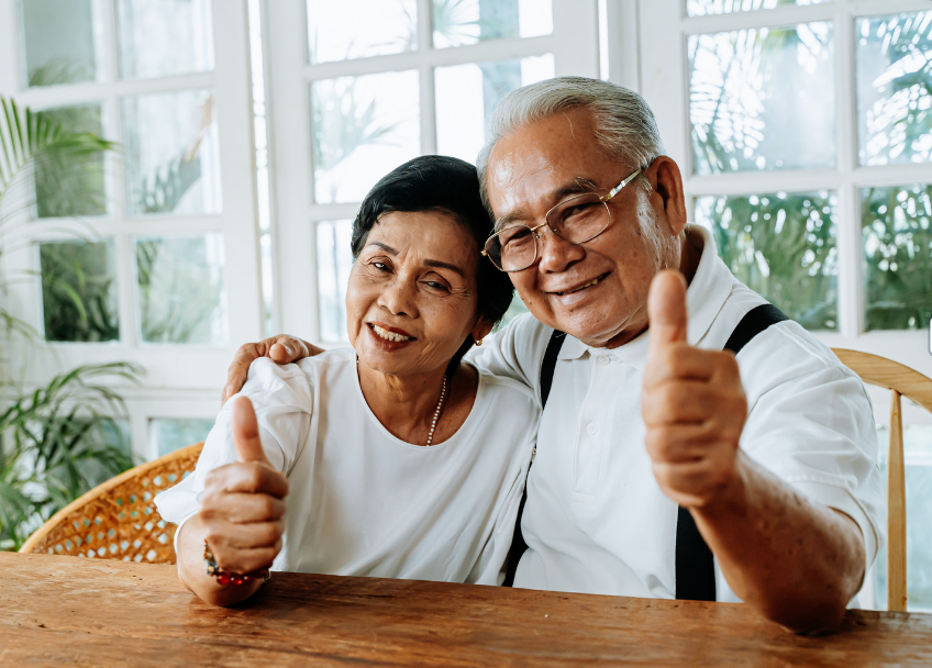 An elderly couple is sitting at a table giving a thumbs up.