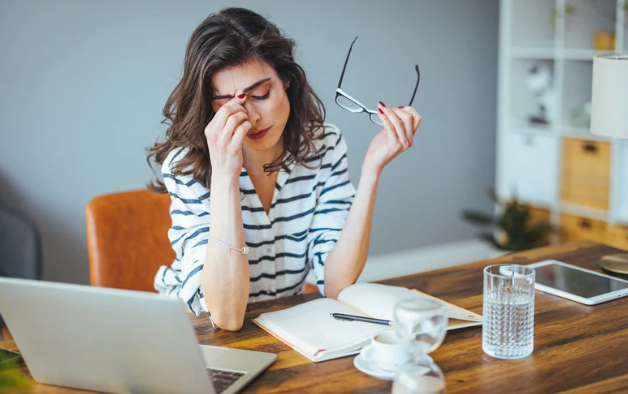 A woman is sitting at a desk with a laptop and rubbing her eyes.