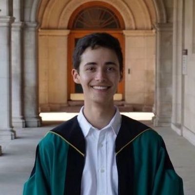 A young man in a graduation cap and gown is smiling in front of a building.