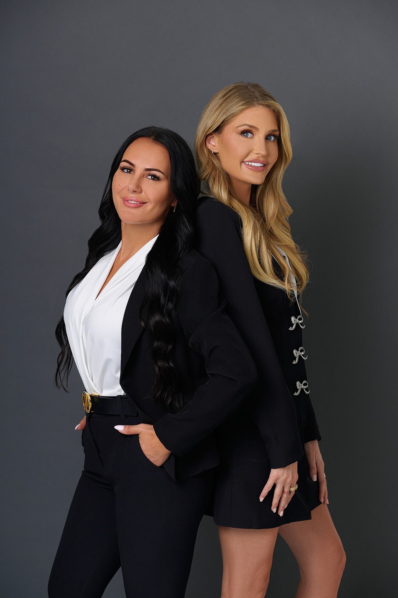 Two women are standing next to each other in front of a gray background.