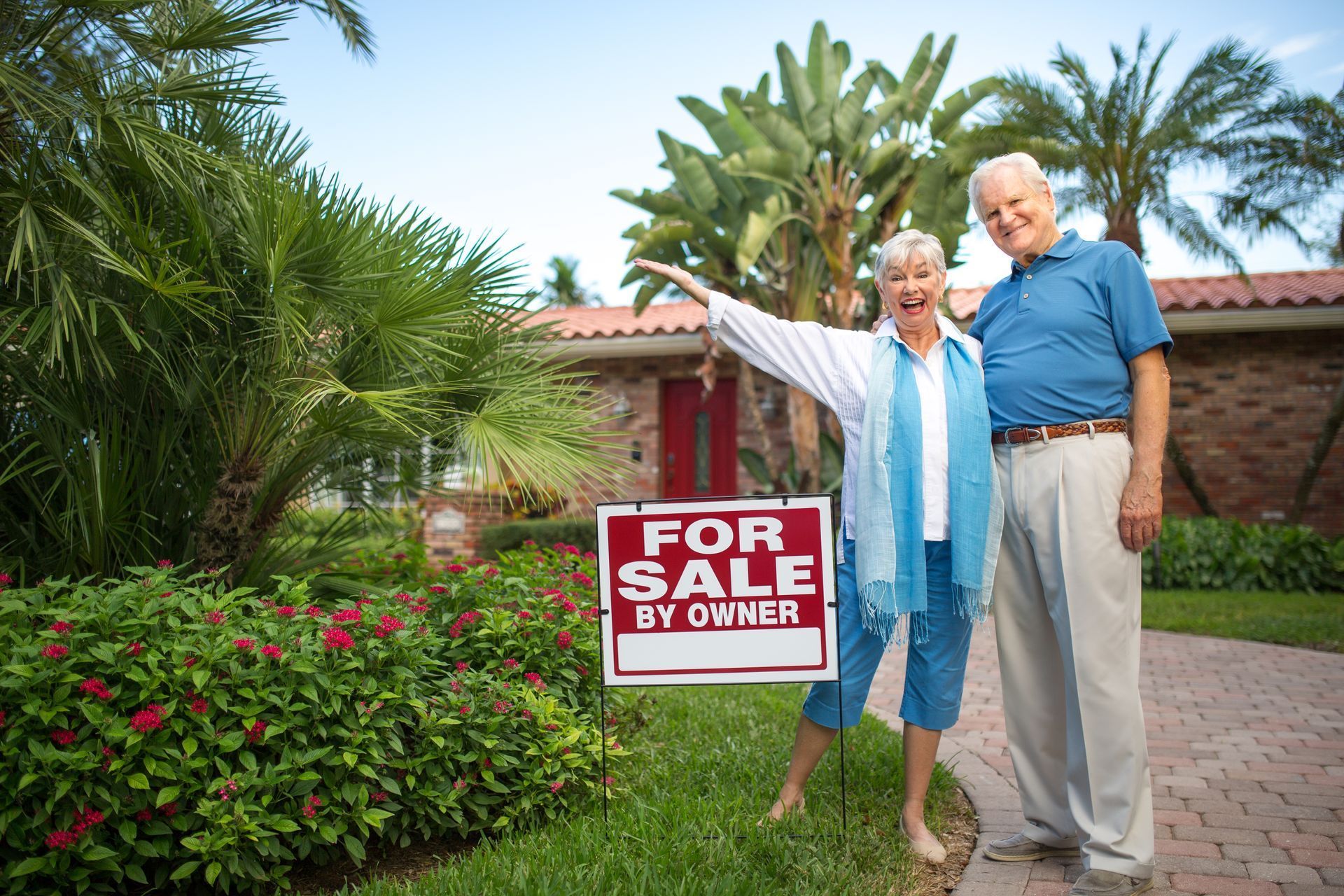 An elderly couple standing in front of a house holding a for sale by owner sign.