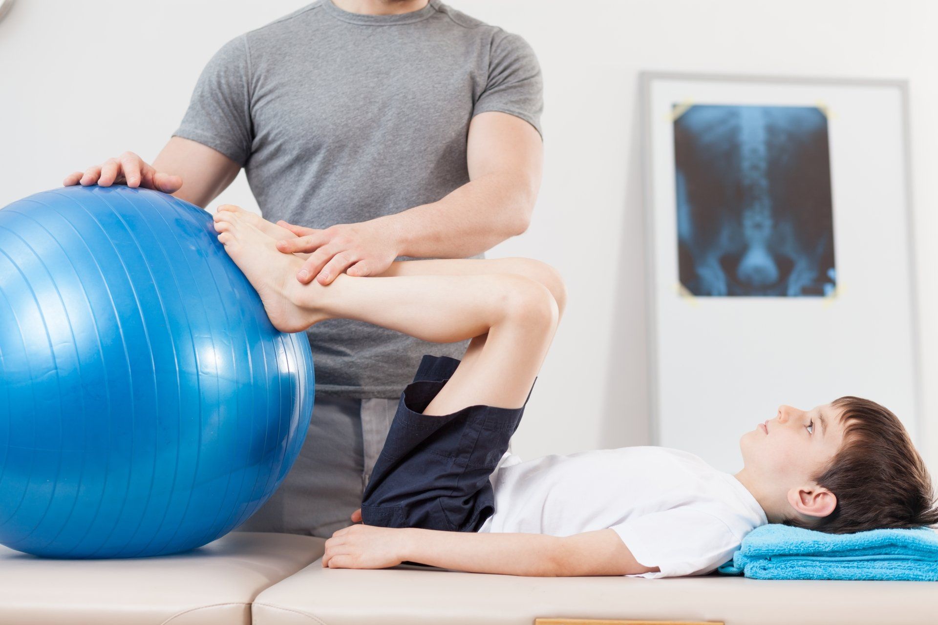 A young boy is laying on a bed while a man holds a blue exercise ball.