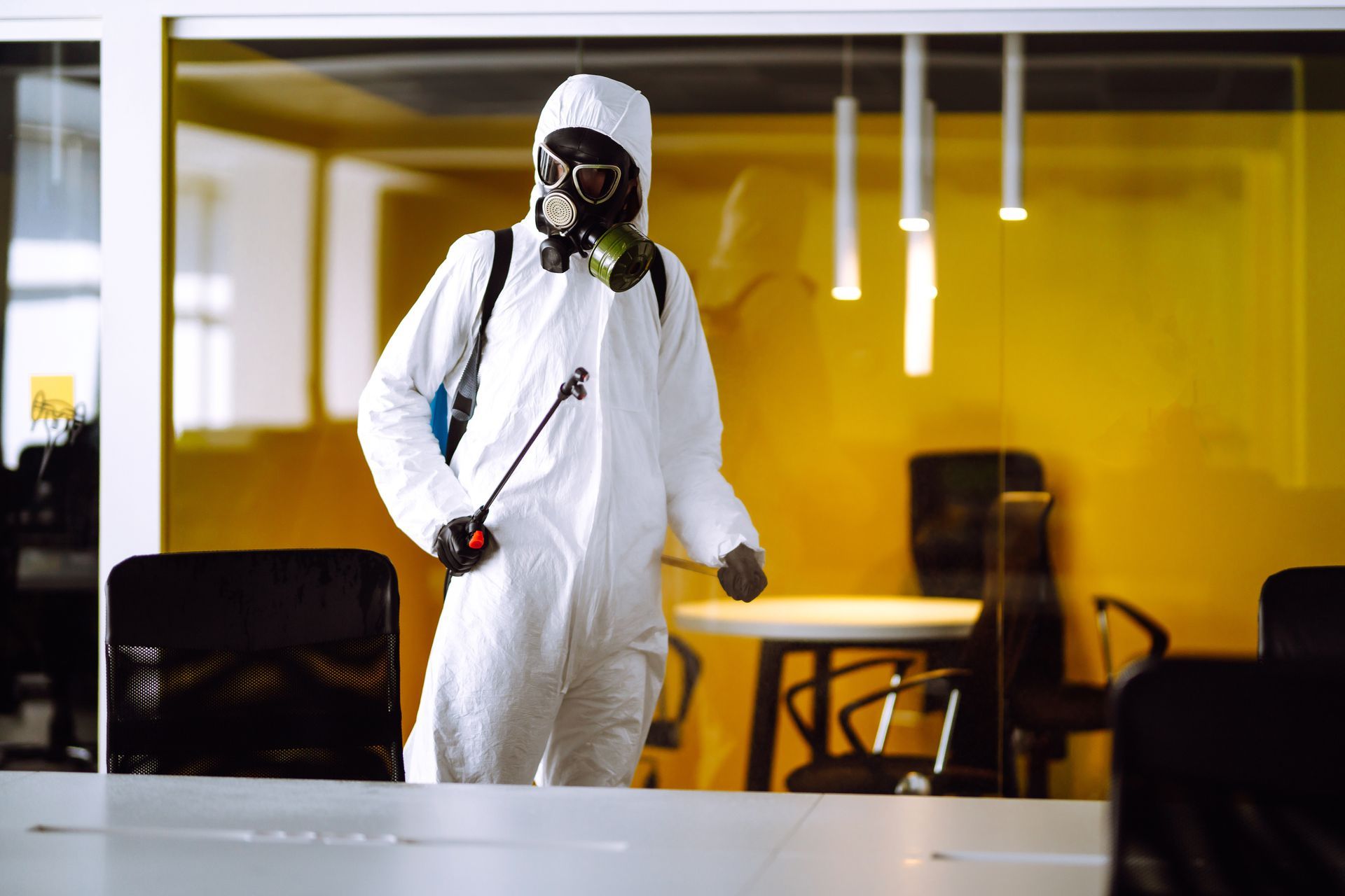 A man in a protective suit and gas mask is disinfecting an office.