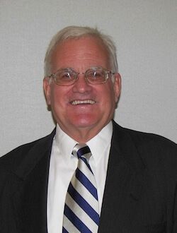 Smiling older man wearing glasses, a suit, white shirt, and blue striped tie.