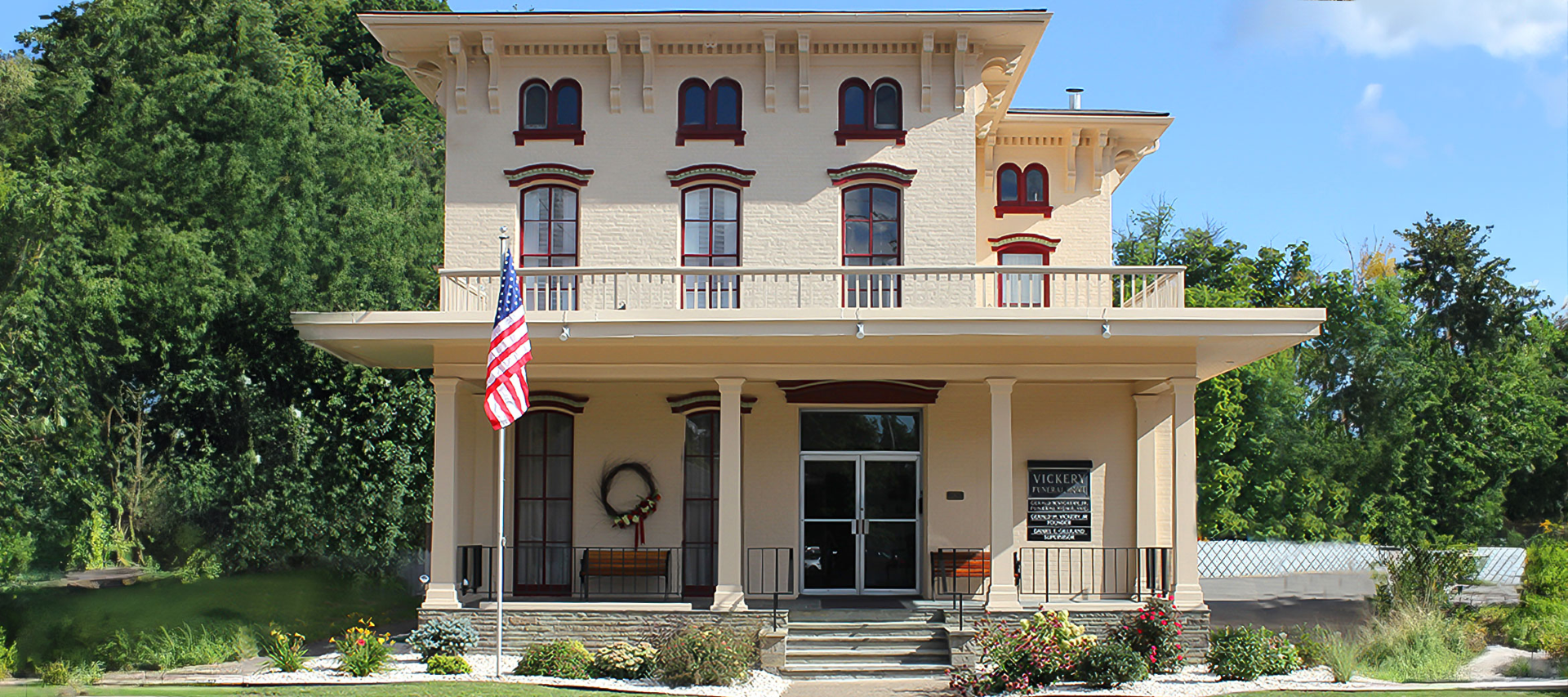 A two-story beige house with a large porch and an American flag in the front yard. Lush green trees frame the house.