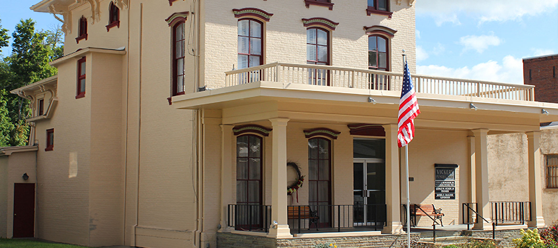 A two-story beige house with a porch and an American flag. The house has dark window frames and a balcony.