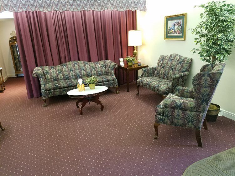A waiting room with floral-patterned furniture, a small table, burgundy carpet and curtains, and a potted plant.