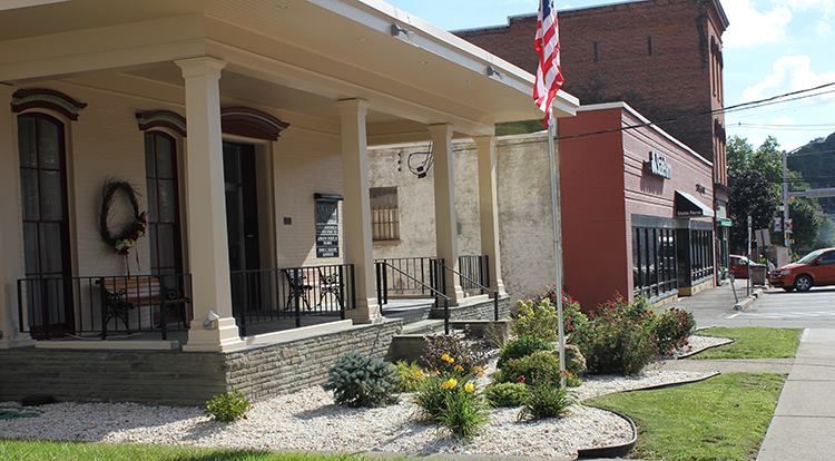 Street view with a light-colored building with porch, American flag, and red brick building. Green plants and white gravel border the sidewalk.