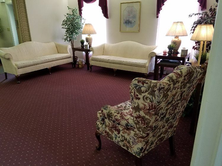 A room with two cream sofas, a floral armchair, and side tables with lamps. Burgundy carpet and white walls.