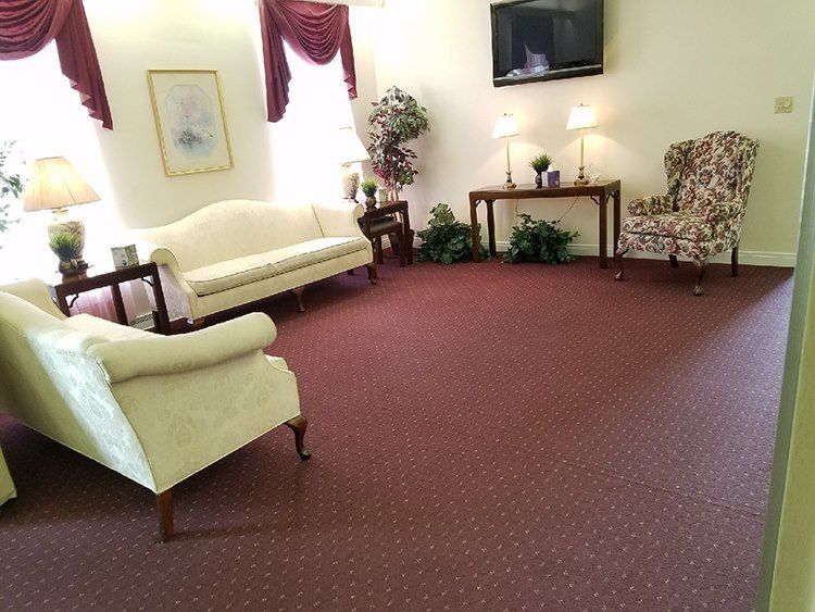 A living room with cream-colored sofas, floral armchair, burgundy carpet, and a mounted TV.