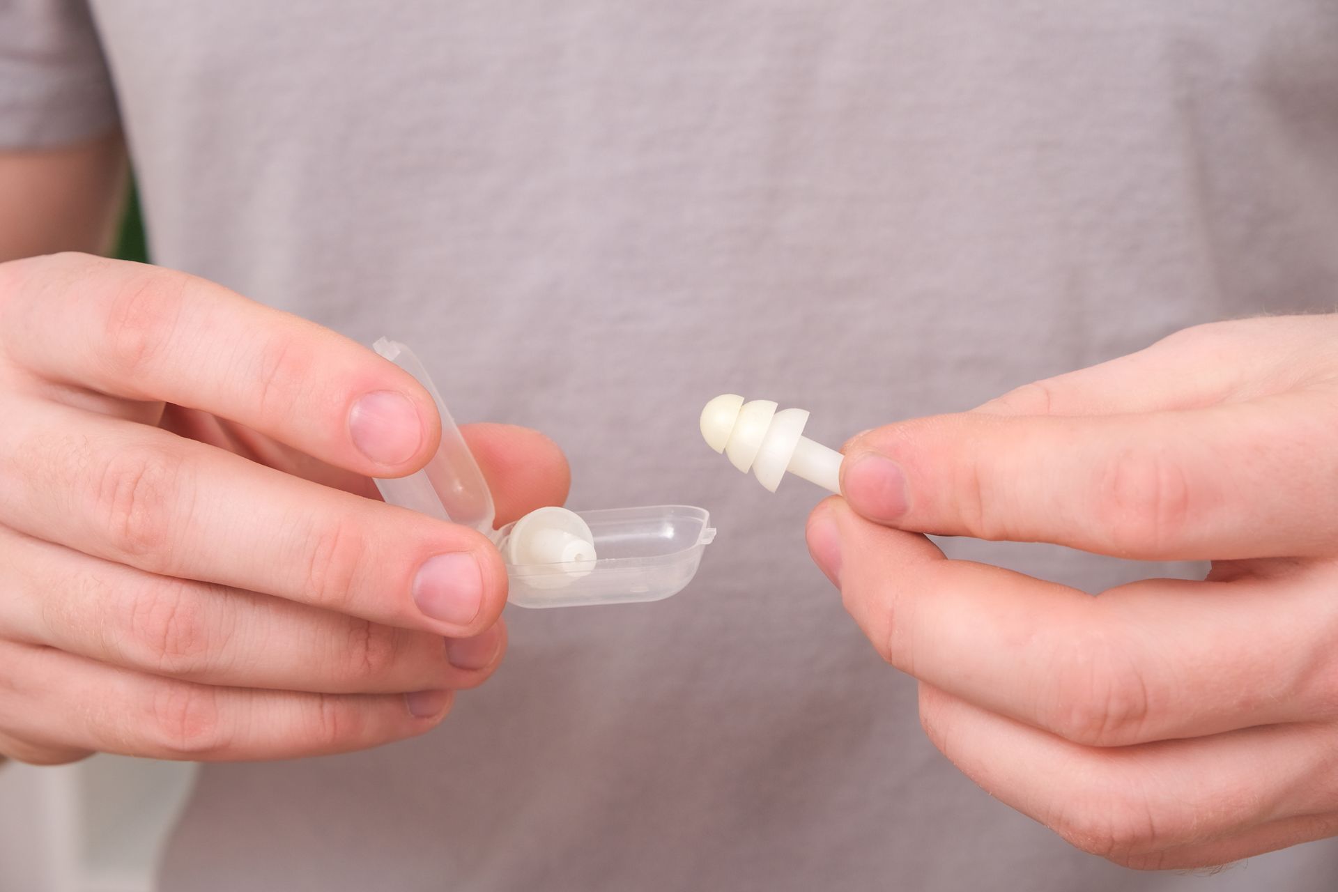 Hands holding a small white earplug and clear case against a plain shirt background