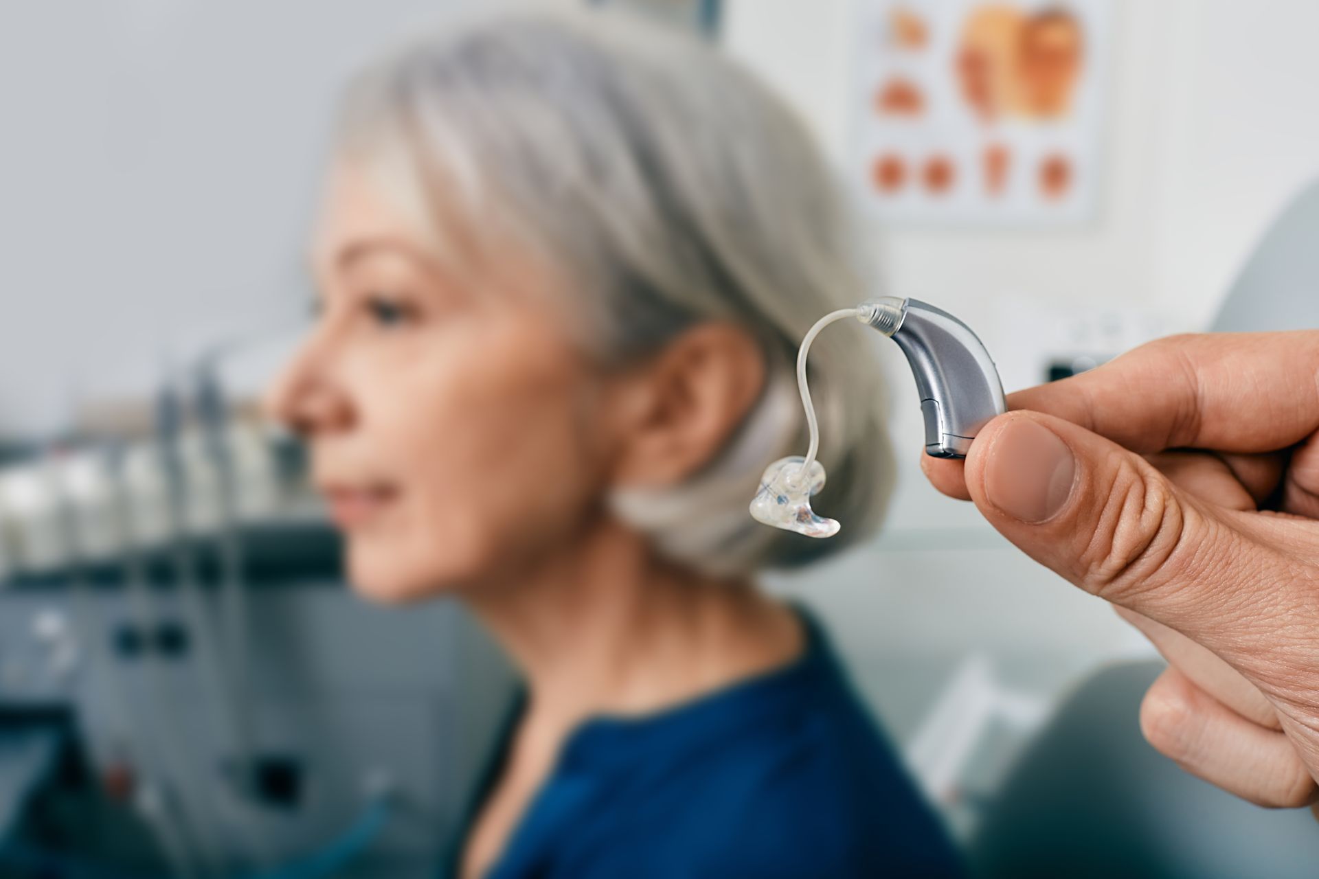 Hearing aid held in foreground with blurred woman wearing one in a clinic setting