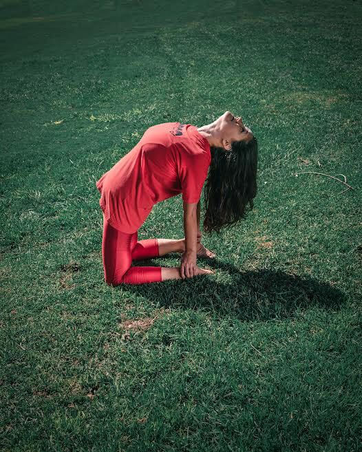 Mujer con ropa deportiva roja haciendo yoga sobre césped verde, con la espalda arqueada y la cabeza erguida.