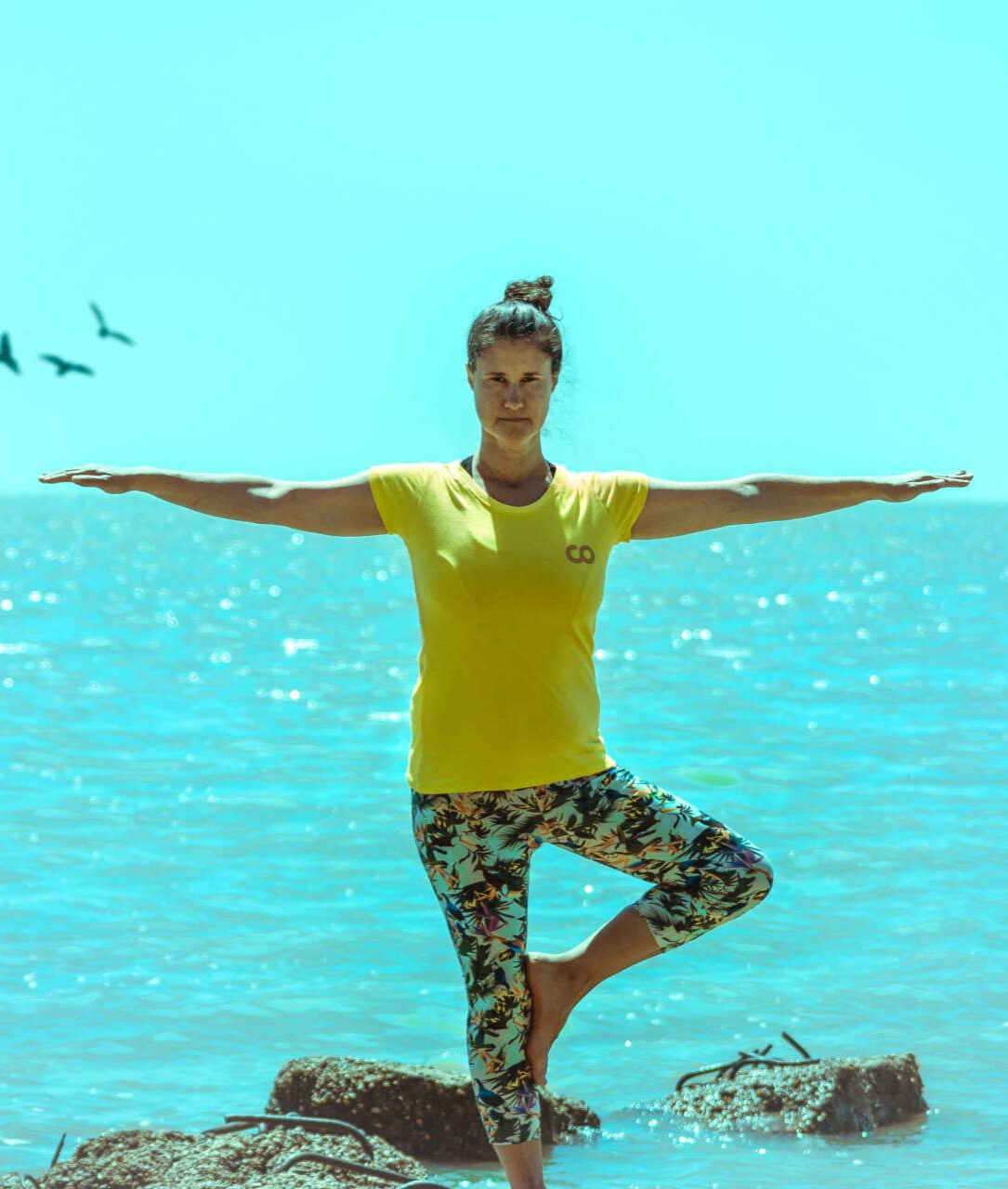 Una mujer con una camisa amarilla y leggings estampados se balancea en una postura de árbol de yoga cerca del océano.