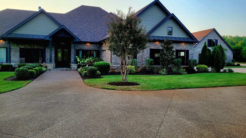Lush green backyard with stone path, patio furniture, and colorful landscaping next to a two-story house under a blue sky.
