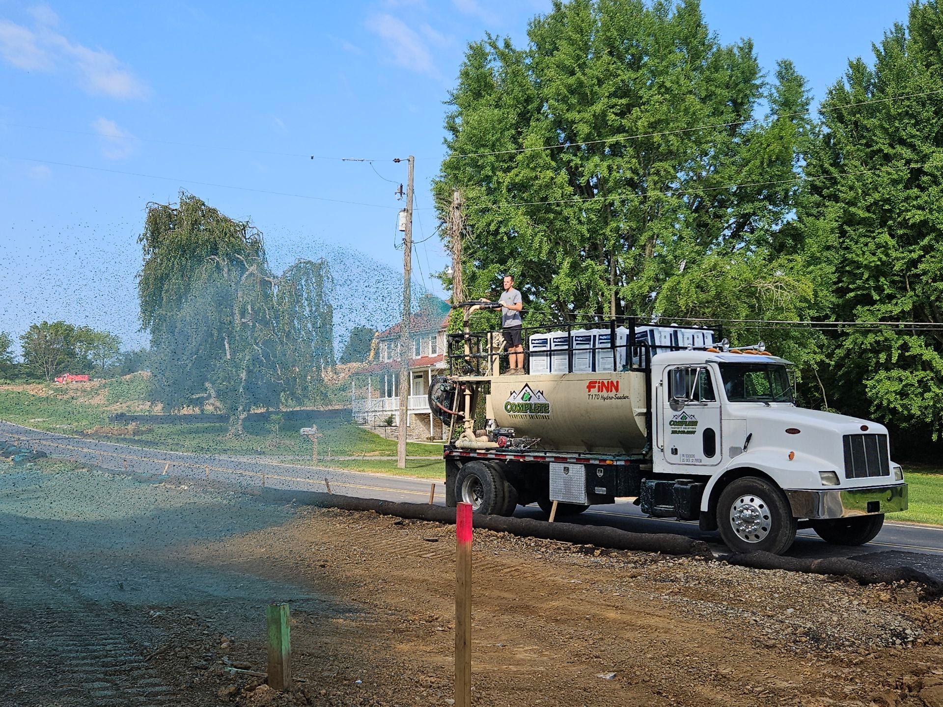 A hydroseeding truck spraying a dirt lawn in Lancaster PA; a worker stands on top. Blue sky, green trees.
