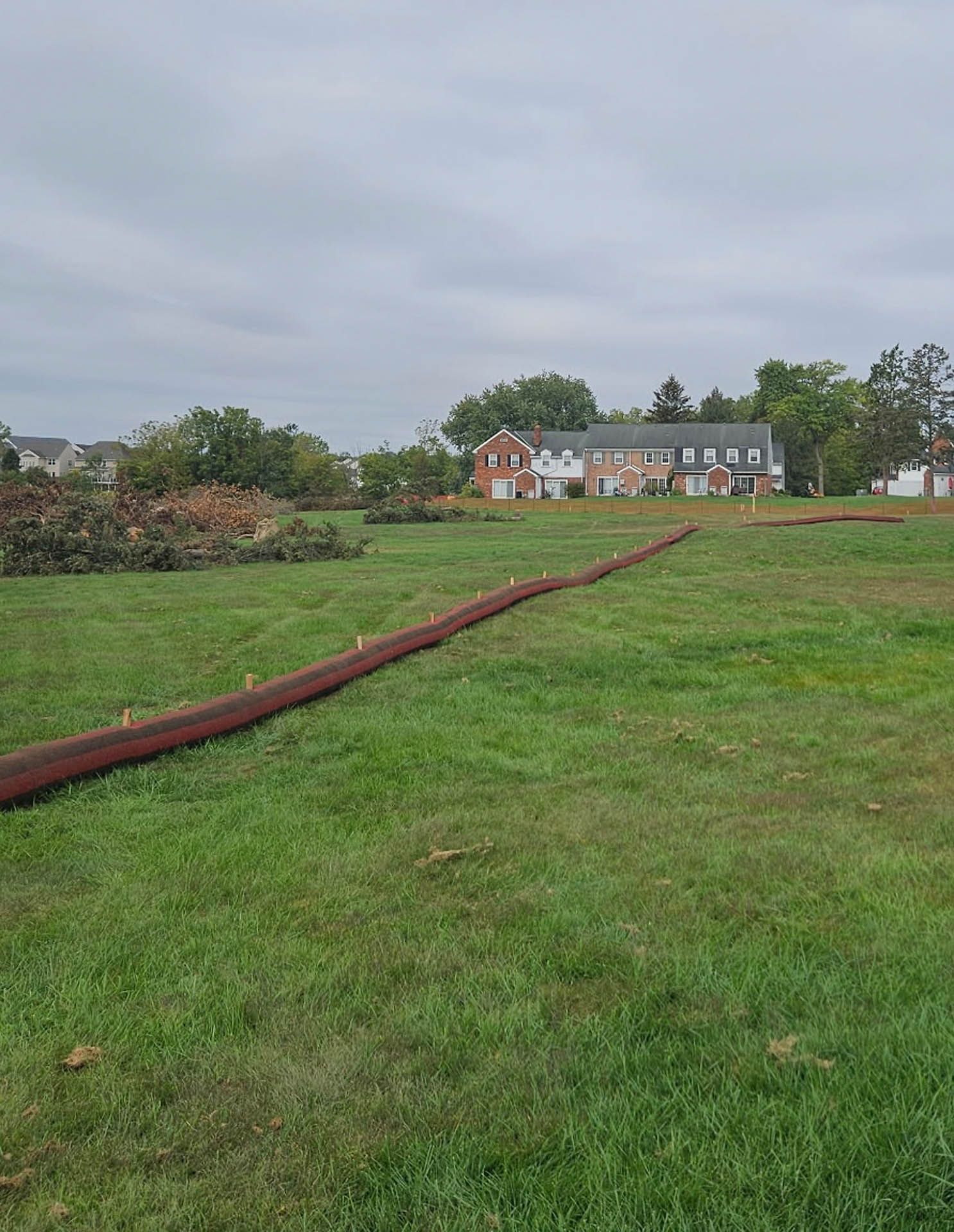 Brown sediment control tube in grassy field, residential houses in the background. Overcast sky.