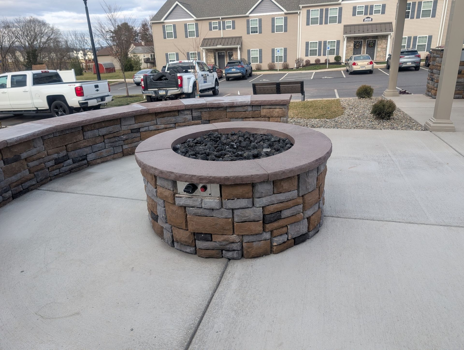 Circular stone fire pit with dark glass, surrounded by a low stone wall, on a concrete patio.