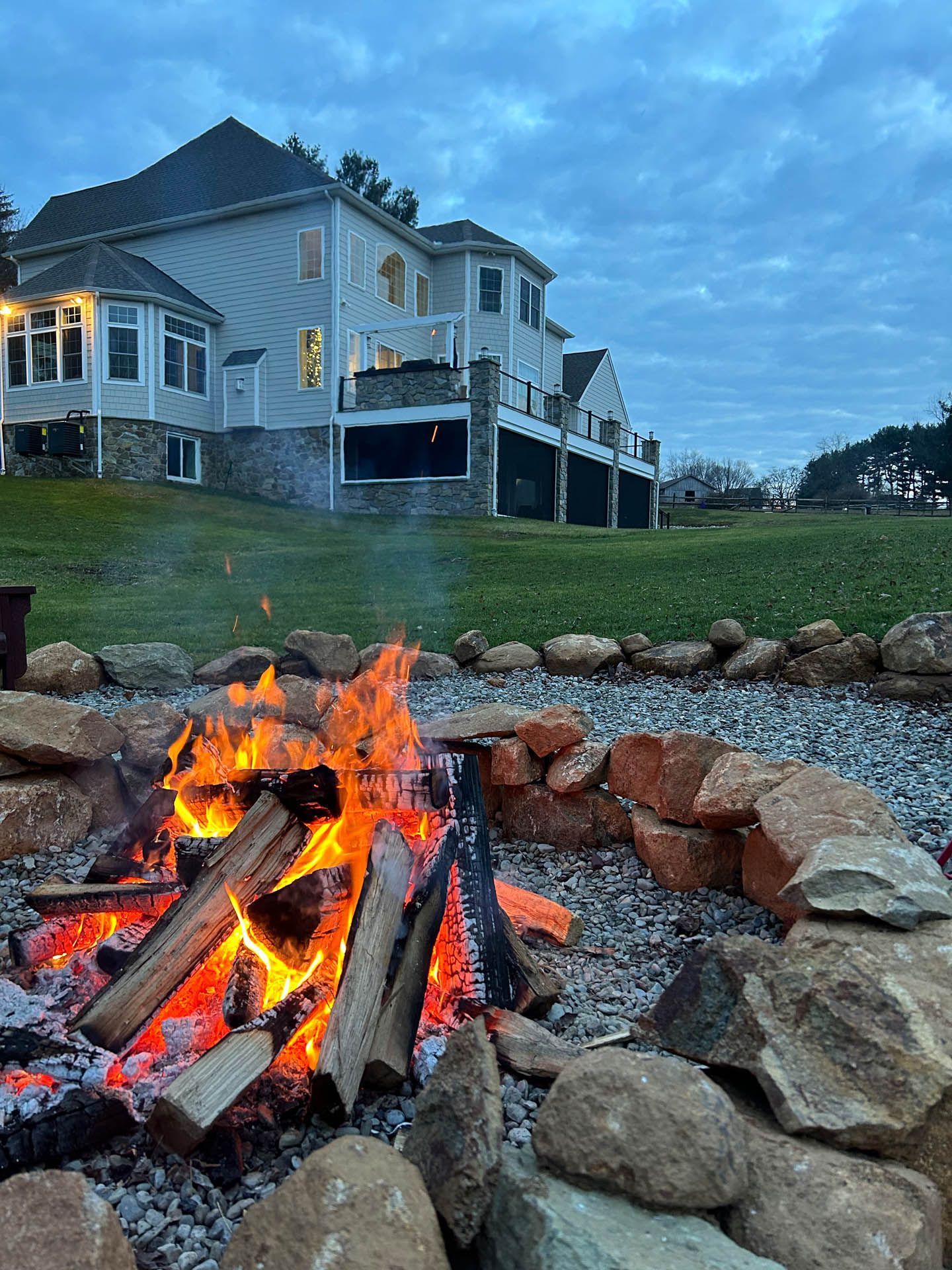 Fire pit with flames, grill, and stacked stones in front of a large house on a green lawn at dusk.