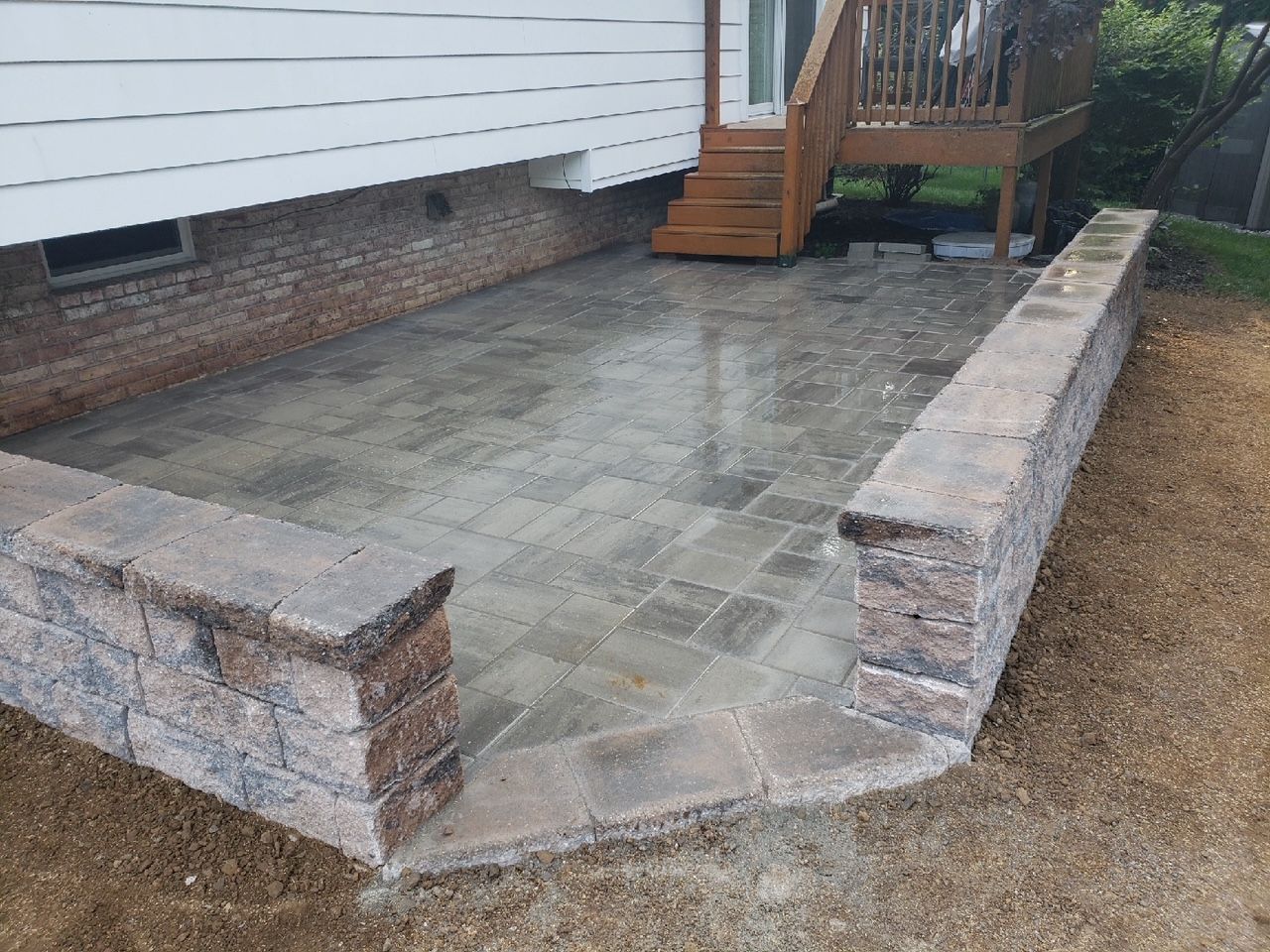 Brick patio with low retaining walls next to a house with a deck and stairs.