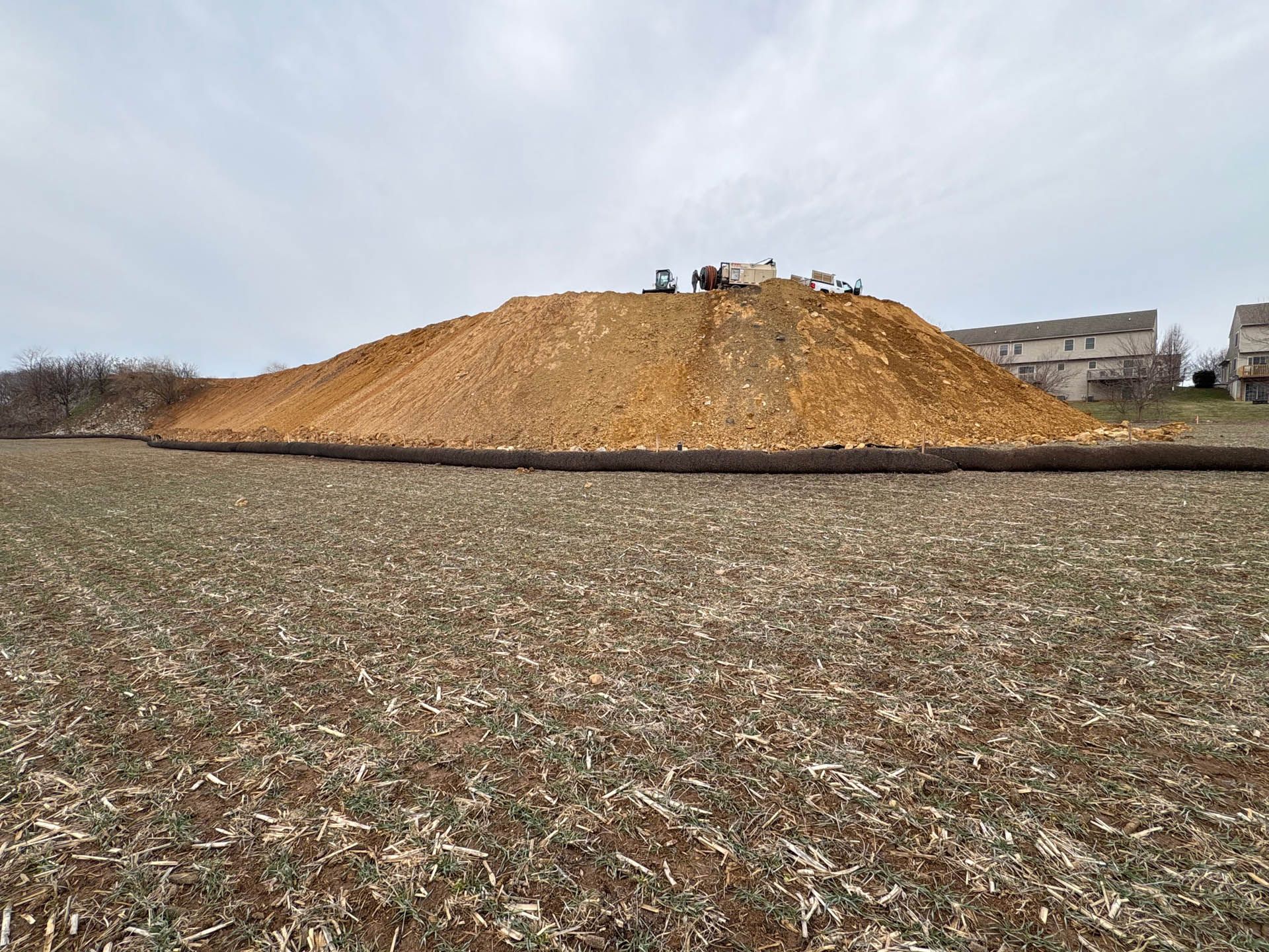 Large pile of wood chips on a field; heavy machinery on top. Cloudy sky, harvested field in foreground, buildings in the background.