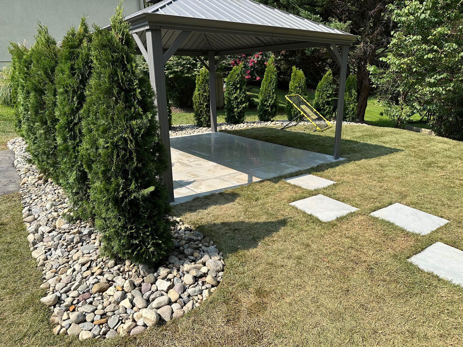Gazebo with stone pathway, next to a row of evergreen trees and rock bed. Sunny day.