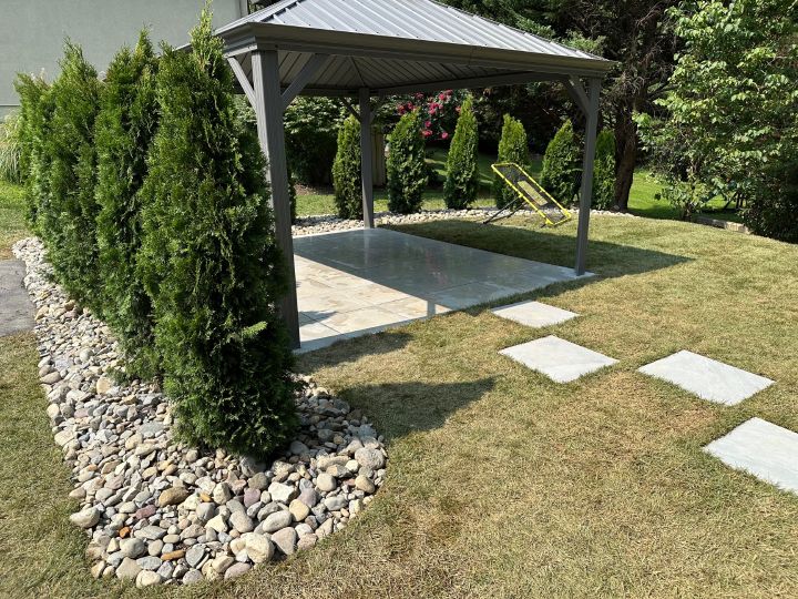 A gazebo with concrete flooring, surrounded by a lawn with stepping stones, and a row of evergreen trees.