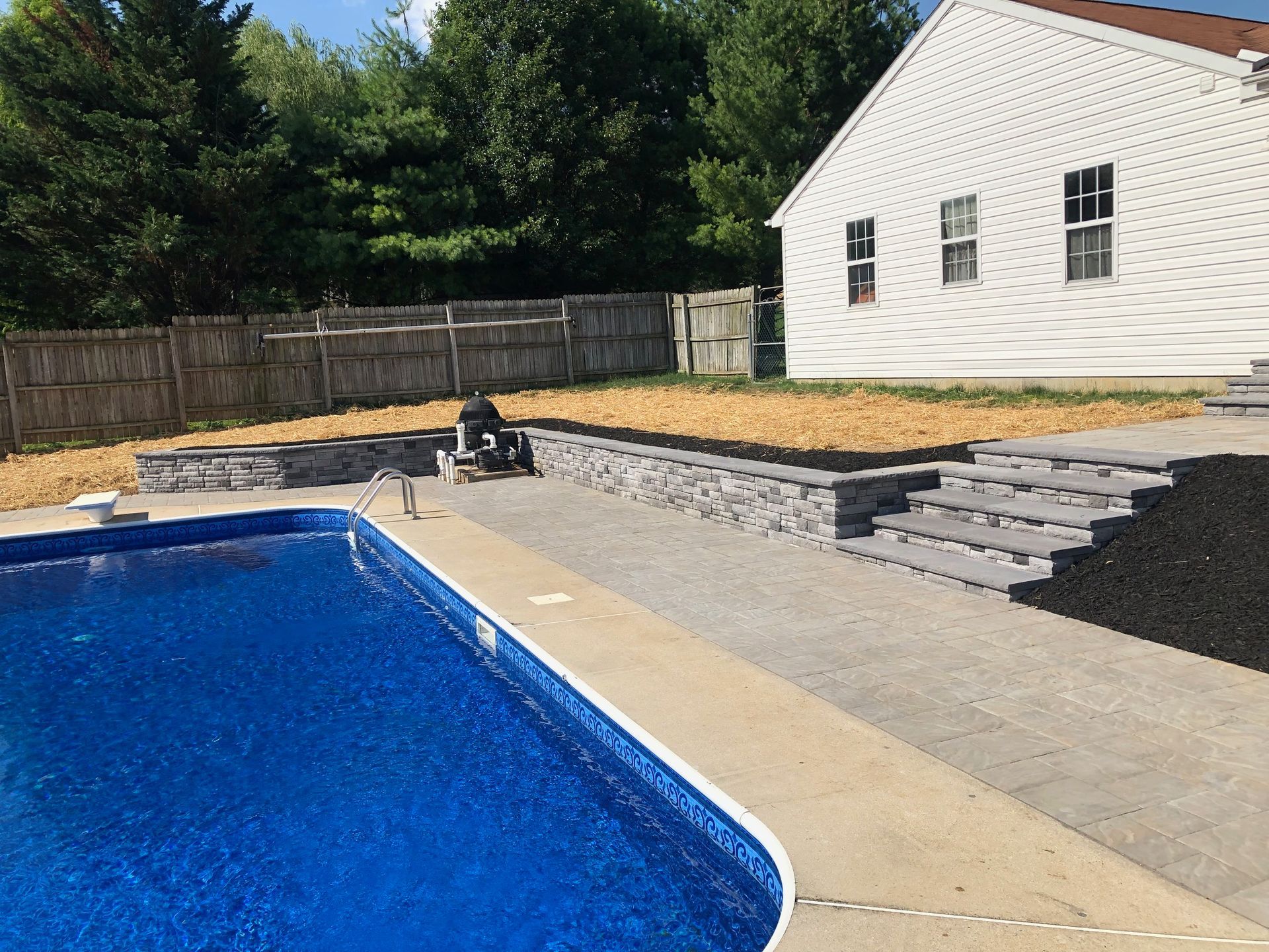 Poolside landscaping with retaining wall, steps, and mulch, next to a white house with a pool filled with blue water.