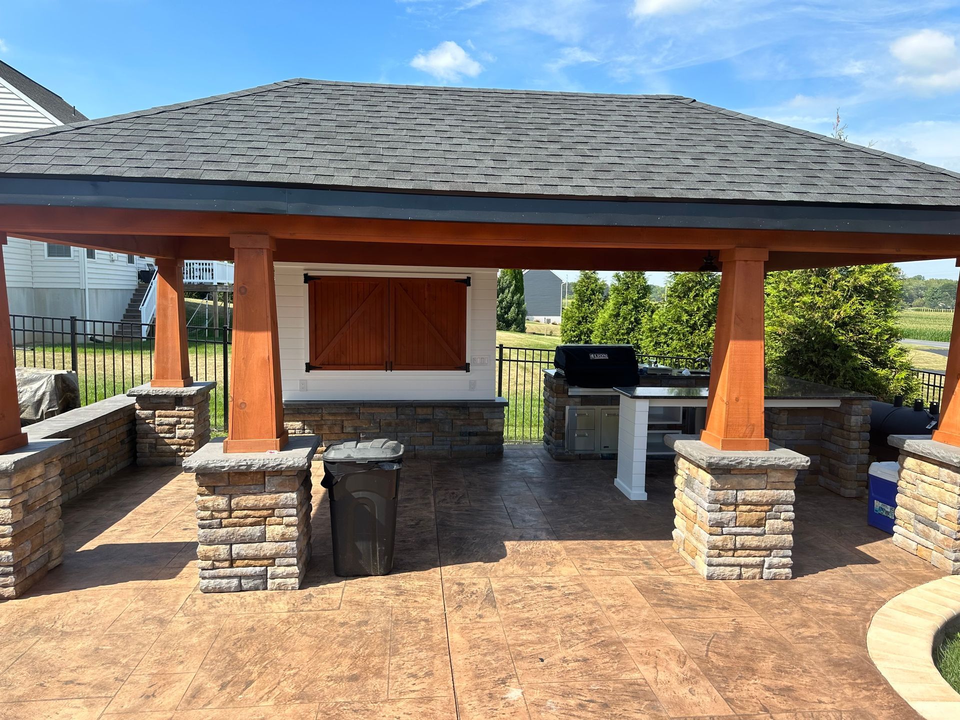 Outdoor kitchen with gazebo, stone pillars, grill, and brown stamped concrete patio under blue sky.