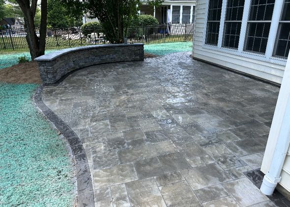 Paver patio with curved stone retaining wall next to a house, bordered by green grass.