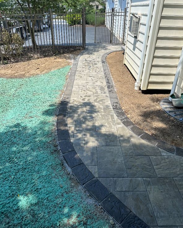 Paver walkway with dark edging curves past a white building and gate, bordered by grass and gravel.