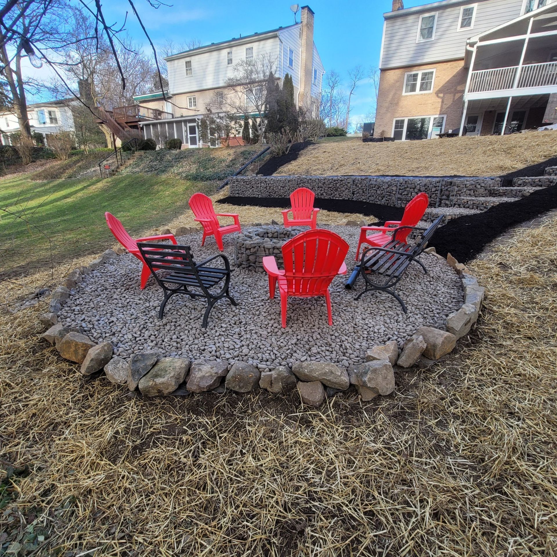 Fire pit with red and black chairs in a backyard with houses on a slope.