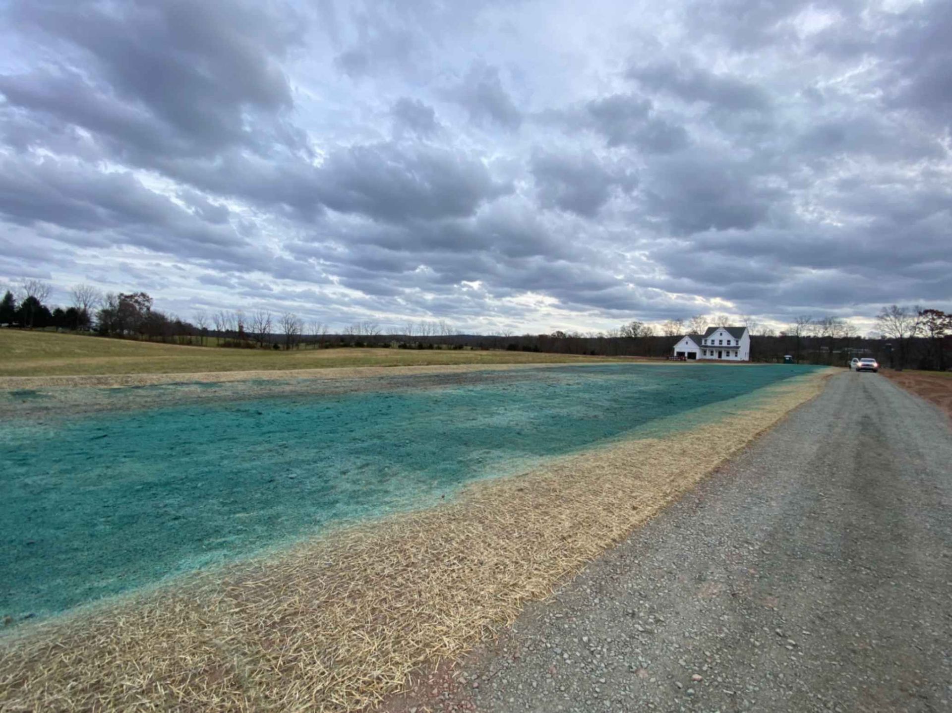 Gravel road next to a field with green-tinted soil. A white house sits in the distance under a cloudy sky.