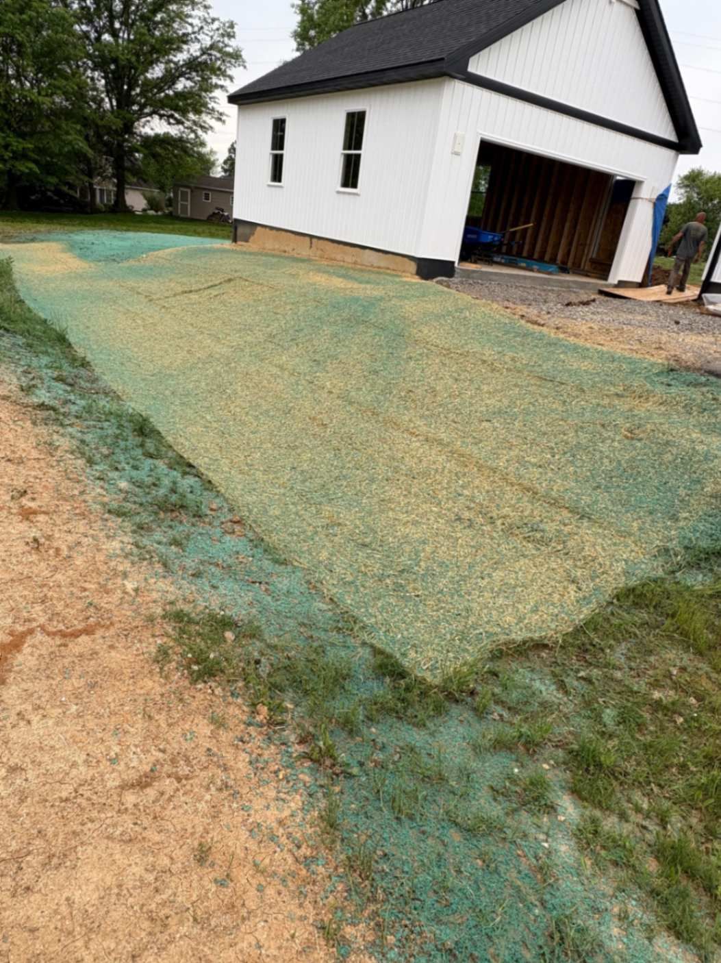 Green seeded mat covers dirt driveway in front of white garage.