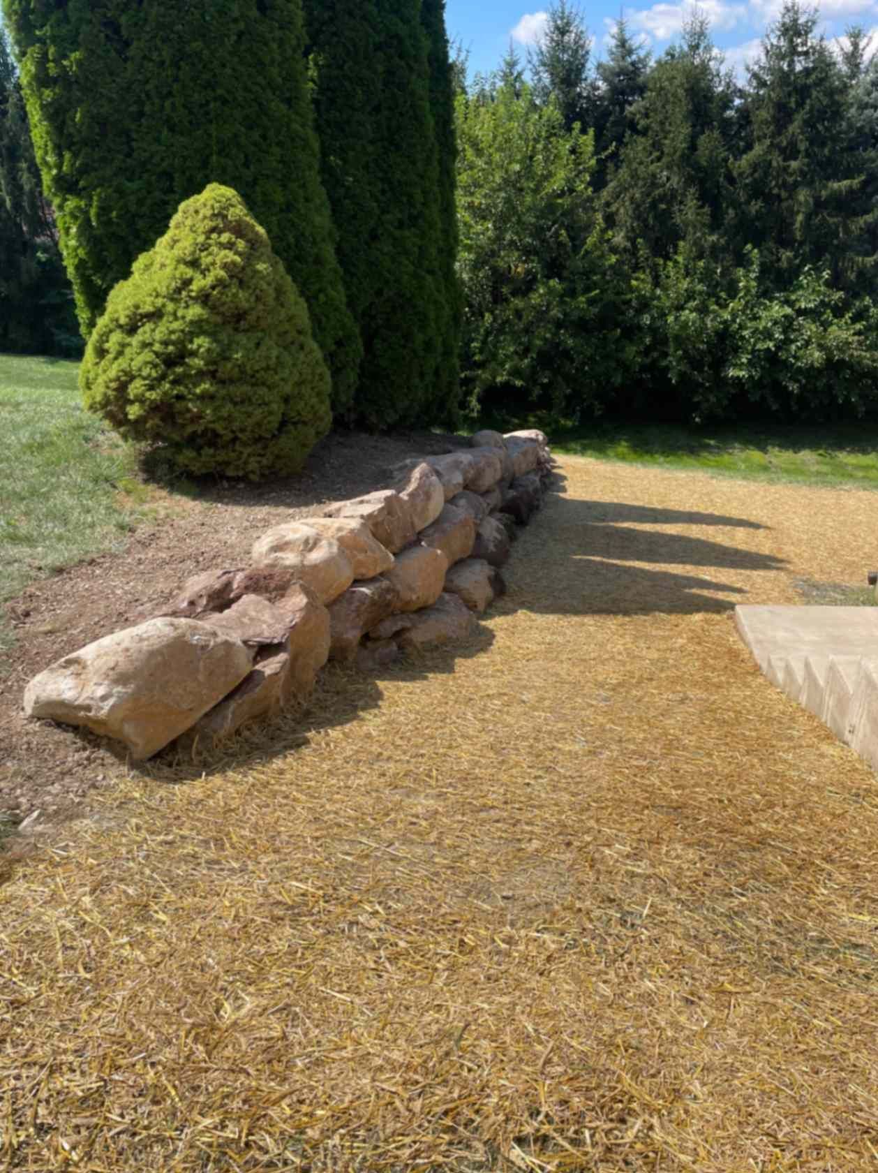 A rock wall bordering a gravel area, beside green bushes and grass.