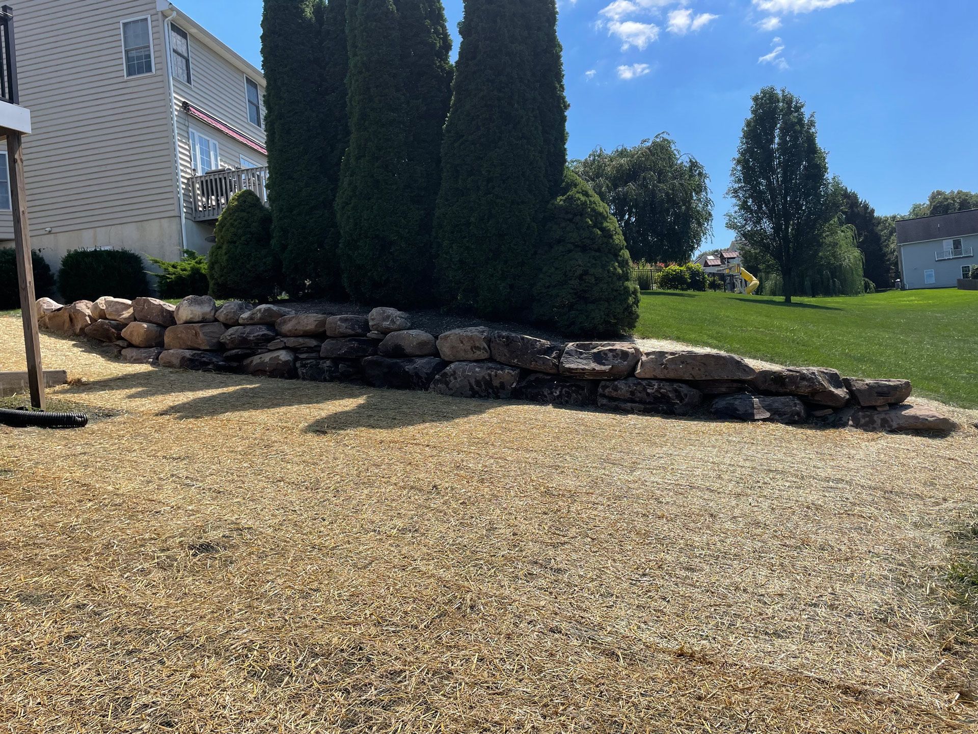 A rock wall bordering a gravel area, beside green bushes and grass.
