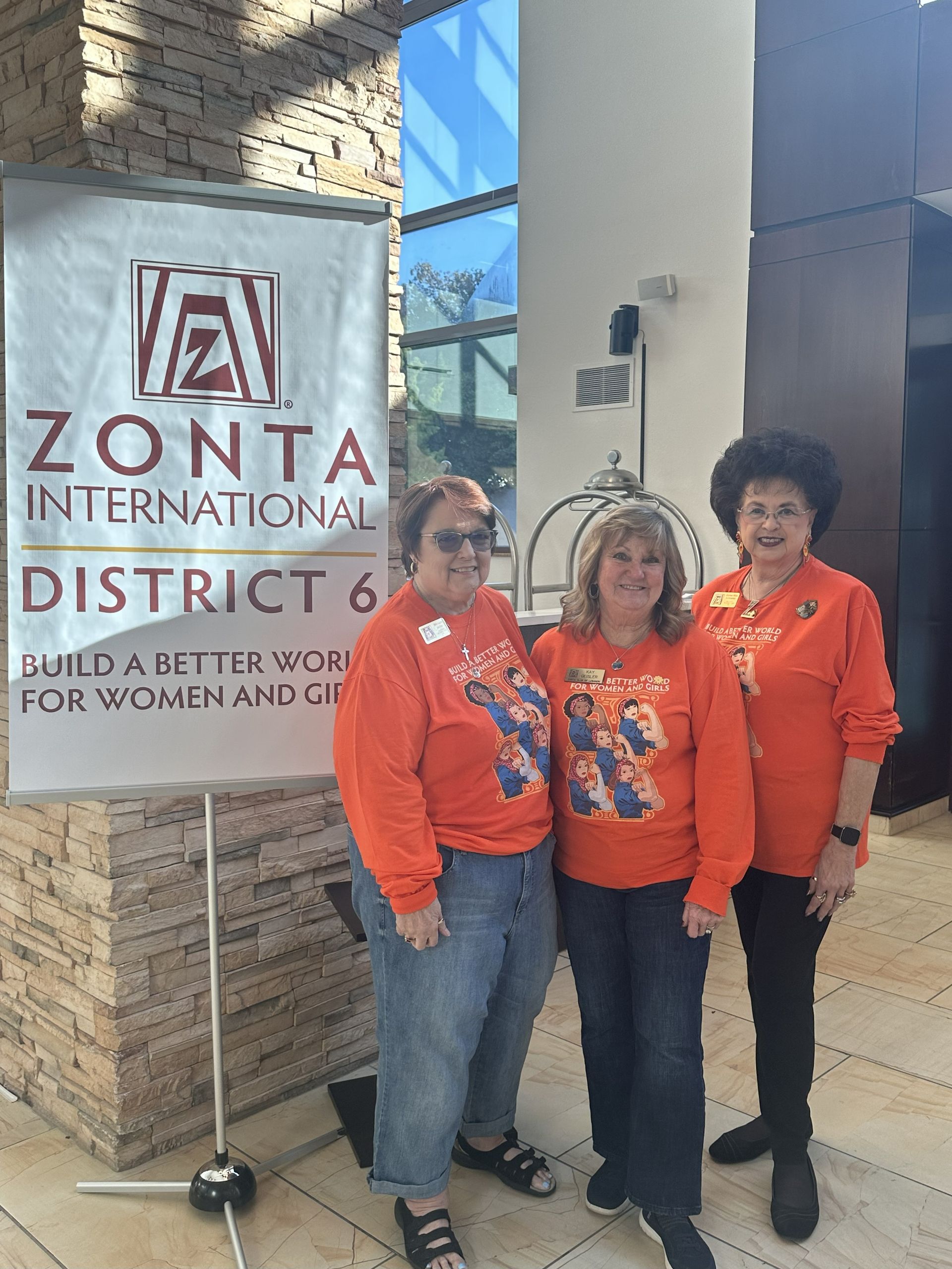 Three women in orange shirts stand by a Zonta International banner in a building.