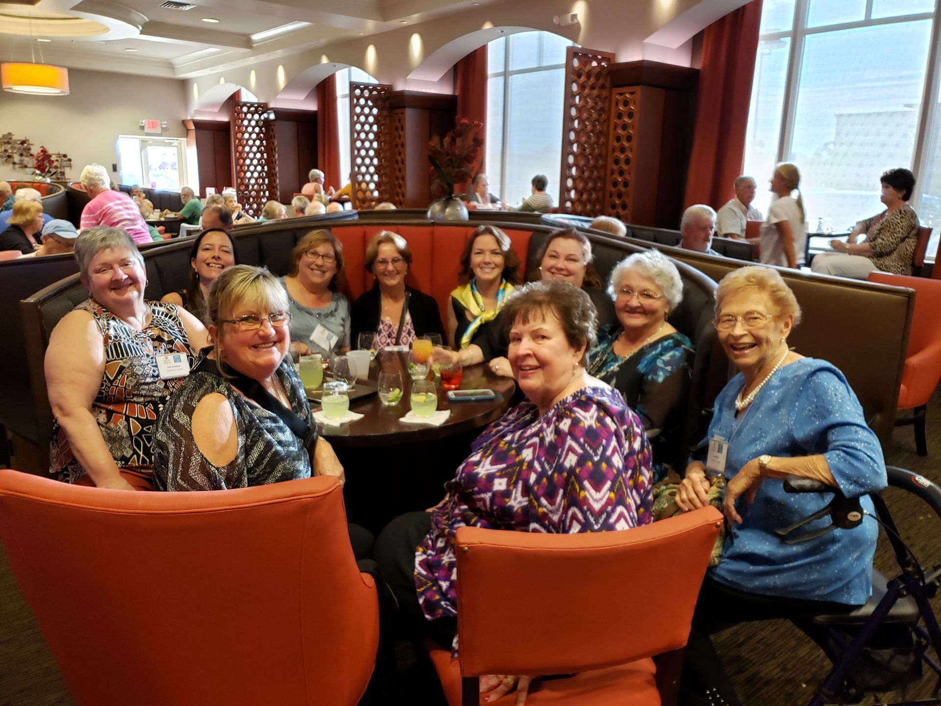 Group of women smiling, seated around a table in a restaurant booth with orange chairs.