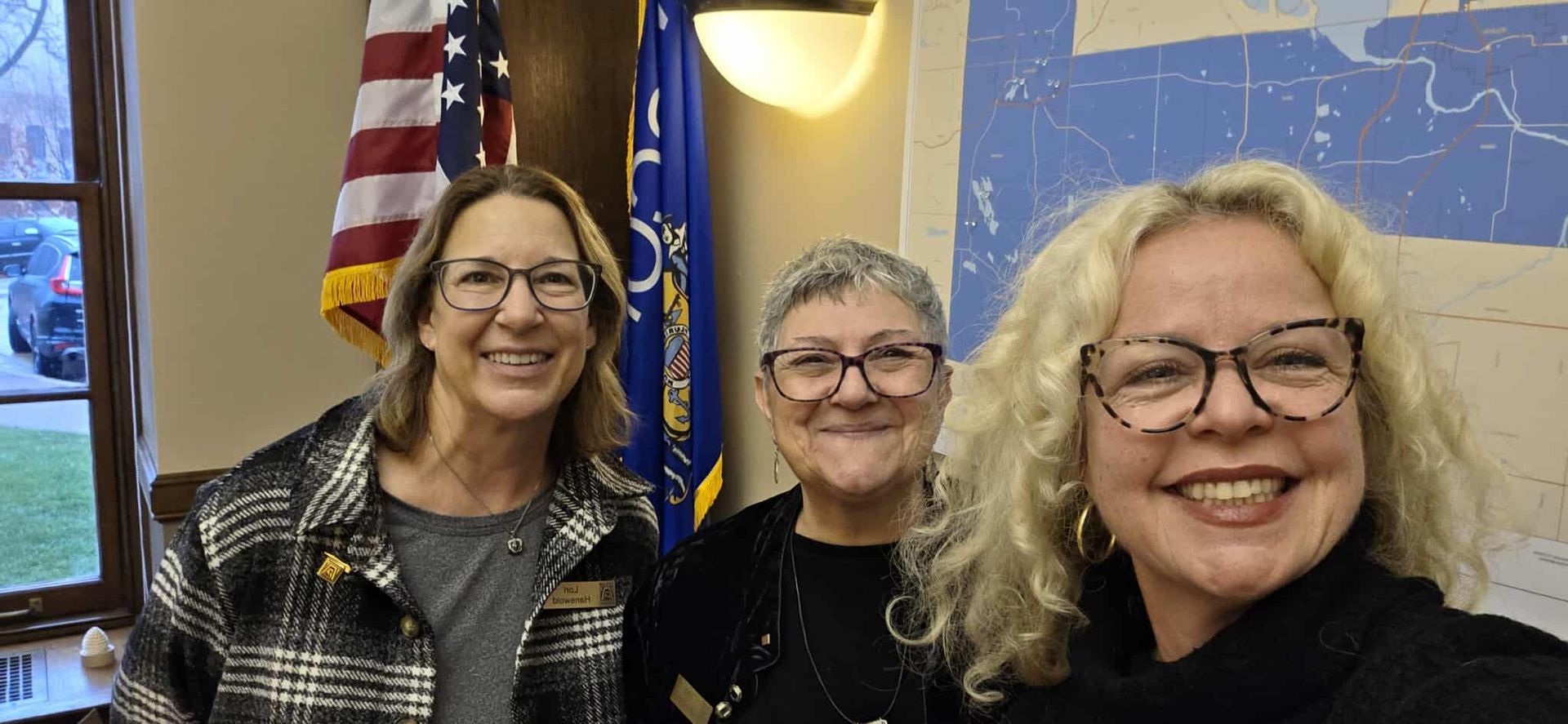 Three people smiling in front of flags and a wall with a mural.