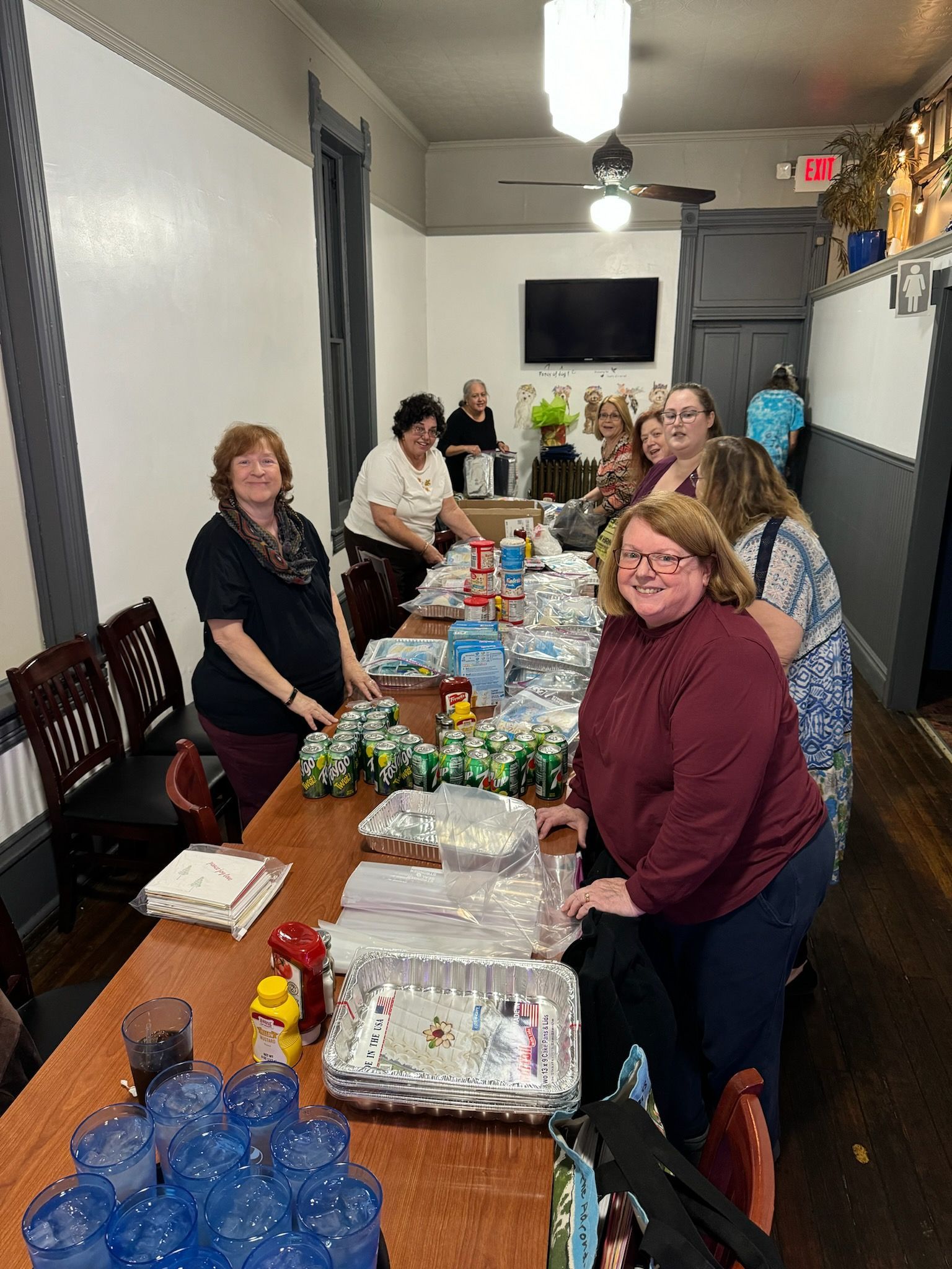 People preparing food at a long table indoors. Canned drinks, bottled water, and prepared meals are visible.