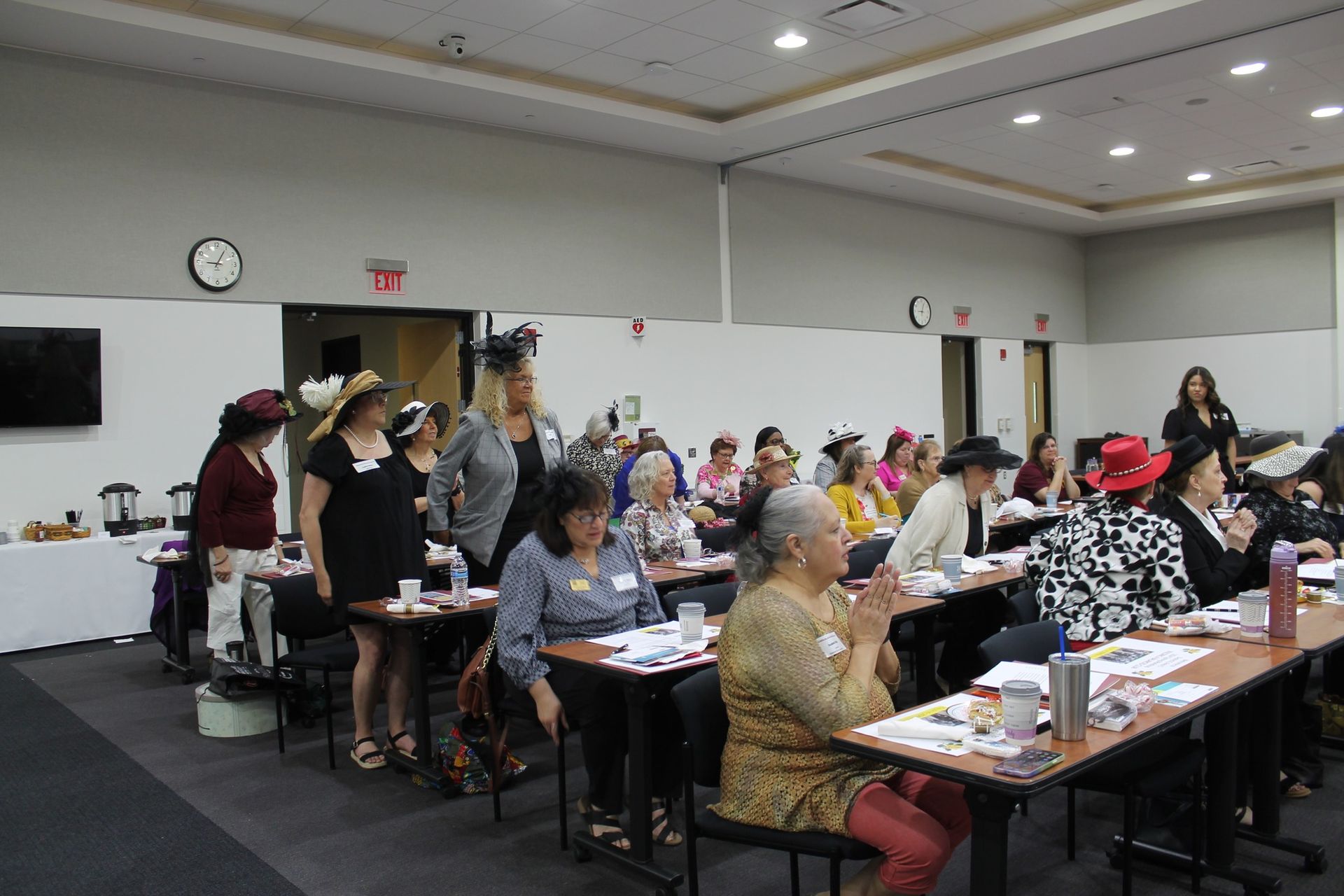 People in a conference room wearing hats; many are seated at tables, others are standing.