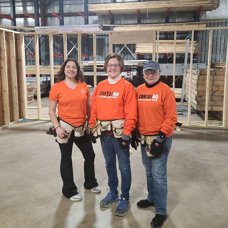 Three women in orange shirts, tool belts, pose inside a warehouse with wooden frames, smiling.