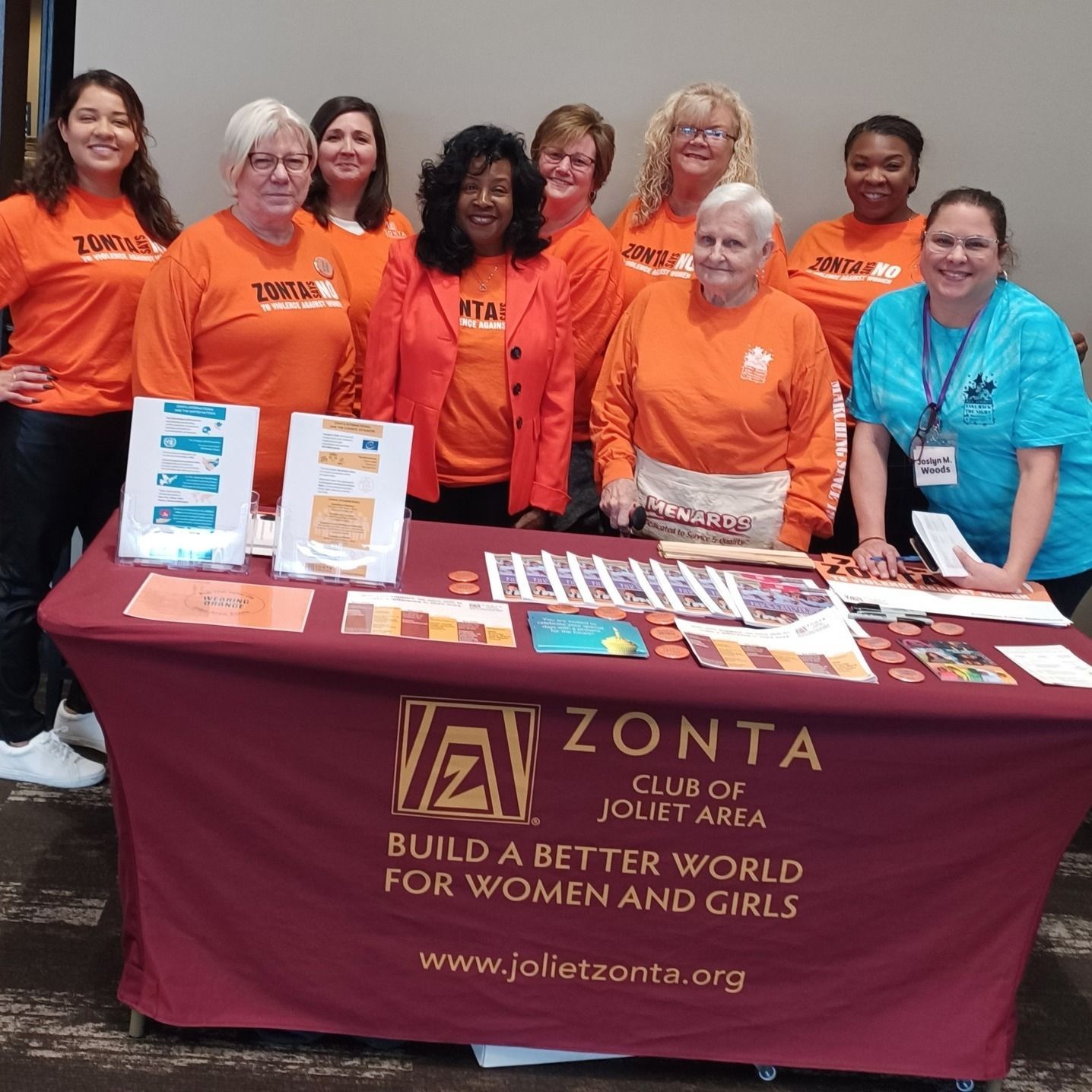 Group of women at a table promoting Zonta Club; they wear orange shirts and stand at a table with brochures.
