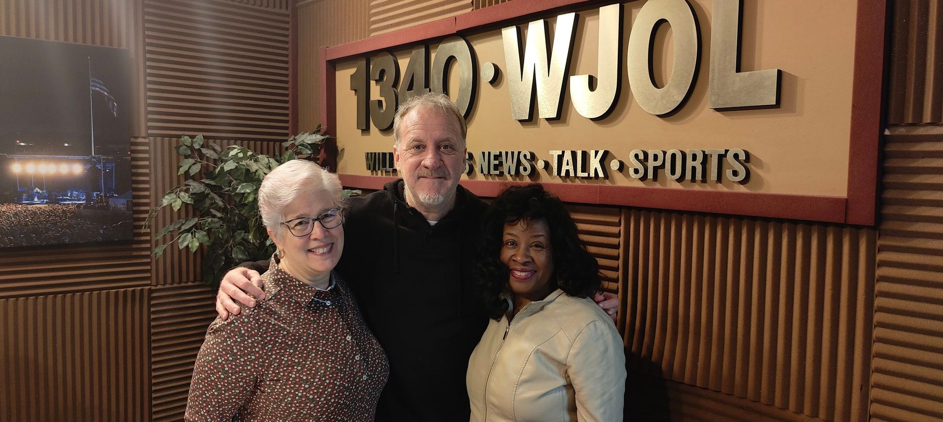 Three people smiling, posing together in front of a radio station sign. The sign reads 1340 WJOI.